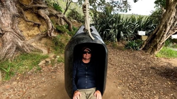 This image shows Adam sitting in a swing that has been made from a huge fender almost the height of him. The fender is black, made of hard plastic and has a rectangle cut out of the front of it big enough for an adult to sit in. The swing is secured to a pōhutukawa tree by multiple knots of a very large diameter rope tied to a purpose built hole in the fender for ropes.