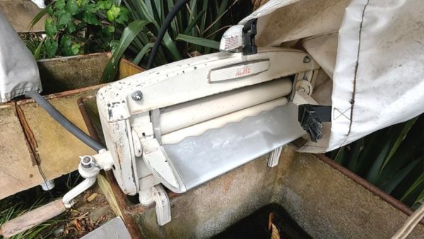 This image shows the manual ACME clothes wringer that is available onshore. It is mounted onto a concrete basin where the water wringed from the clothes can be collected. Once finished with the wringer, there is a white cover that keeps it protected from the elements.