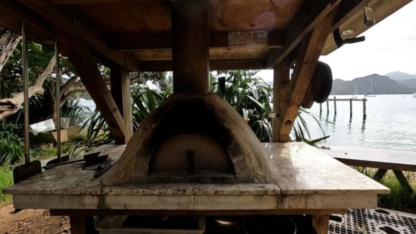 This image shows a pizza oven that is available for use onshore. It is mounted on a table, waist height, and the table has a roof where the chimney for the pizza oven exits. It has a door on the front and smoke residue around the door from being used. In the background you can see the anchorage with the tidal frames. There are also cooking utensils available for use that hang on the table roof around the pizza oven.