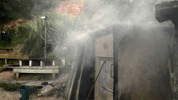 This image shows the smoker where people can smoke their catch of the day. In this image, the smoker is being used and a lot of smoke is escaping the closed doors. The smoker is two ovens wide and is stands approximately 1.5-2 meters tall. It is made of some sort of metal and is well stained from all the smoke that has escaped. It has two doors on the front of it which are closed when in use and there is a fire poker leaning on one of the doors. In the background you can see the barbeque area.