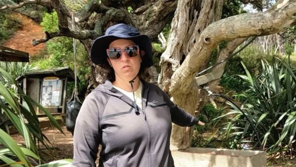 In this image, Vicki is pointing to the concrete washing basins onshore where boaties can wash their laundry. Next to the concrete washing basins is a very big pōhutukawa tree which provides the basin with shade.