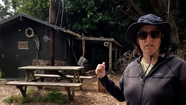 In this image, Vicki is pointing to the bath house which is behind her. In front of the bath house is a wooden table with bench seats attached to each side of it. Like a table you would find at a park. The wash house is green in colour with a tin roof. It has an open wooden structure on the side of it where wood can be stacked for use in the woodfire. It also has a fish, a life ring, and a piece of wood as ornaments hanging on the front of it. Just in front of the bath house but behind the table and chair set you can see the rusty chimney for the woodfire.